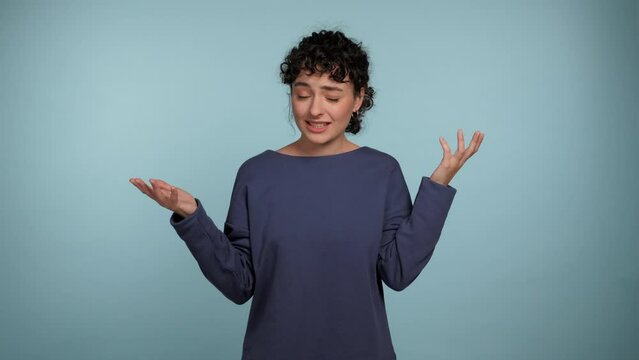 Portrait beautiful confused curly woman spreads hands says what looks camera. Upset female wearing blue sweater on isolated light blue background with copy space. People emotions lifestyle concept