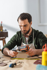 Bearded craftsman in apron holding electric screwdriver in workshop.