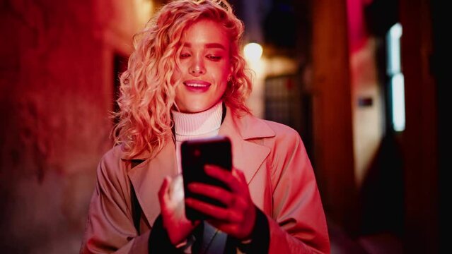 Attractive Female Tourist Walk Down City Street With Red Neon Lights While Holding Cellphone Which Illuminates Her Face In Night.Millennial Young Woman Typing Text Message In Chat Via Mobile Phone App