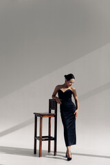 Young woman poses standing near chair in long black dress
