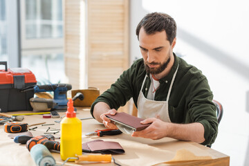 Bearded carpenter in apron holding sandpaper near tools on table in workshop.