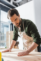 Bearded craftsman in apron touching wooden board in workshop.