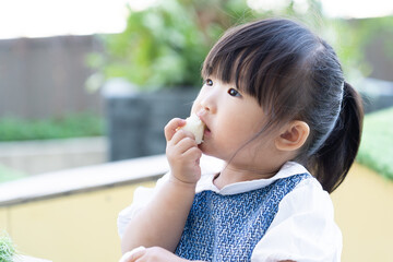 A cute little girl with bangs hair eating snack with her hands while playing outdoor in public park, a kid take some food into mouth at school yard during summer sunny day
