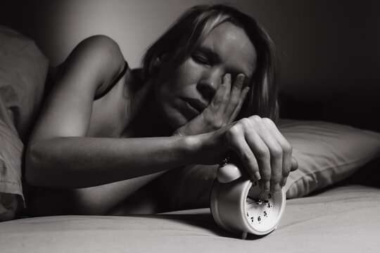 Irritation of a woman in the morning from the alarm clock. Poor sleep, violation of the regimen, lack of sleep concept. A middle-aged woman turns off the alarm clock. Black and white image.
