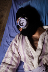 Thief of time, procrastination concept. A man in a balaclava lies and sleeps with an alarm clock on...