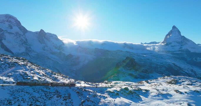 Aerial drone footage of Gornergrat bahn railway running towards summit station with Matterhorn mountain peak background in Zermatt on a sunny winter day. Swiss Alps, Switzerland travel journey trip.