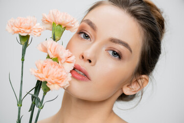 young woman with perfect skin and natural makeup looking at camera near fresh carnations isolated on grey.