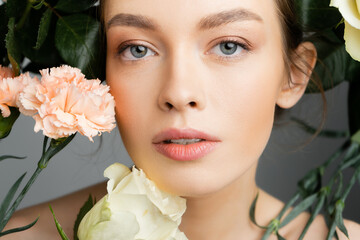 portrait of young woman with natural makeup and perfect skin looking at camera near fresh flowers isolated on grey.