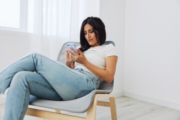 Woman sitting on a chair with phone in hand in a new apartment, communicating online in messengers and social media over the Internet