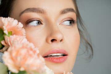 Close up view of woman with natural visage looking away near blurred carnations isolated on grey.