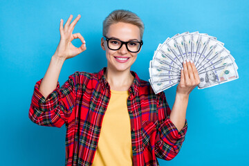 Portrait of positive cheerful woman with short hairdo wear checkered shirt hold dollars showing okey isolated on blue color background