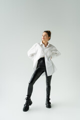 full length of woman in white oversize shirt and black tight pants posing with hands on waist on grey background.