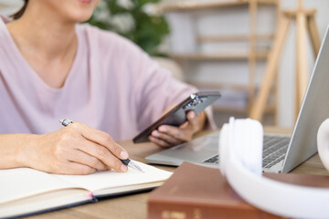 Fototapeta premium Girl college student using smartphone watching video course, zoom calling making notes in workbook sit in living room. Asian woman hold phone online learning in mobile app.
