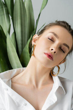 Portrait Of Sensual Woman In White Shirt And Hoop Earrings Posing With Closed Eyes Near Blurred Plant Isolated On Grey.