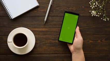 Composition with a hand holding a smartphone with a green screen, clean note, pen, a cup of coffee,...