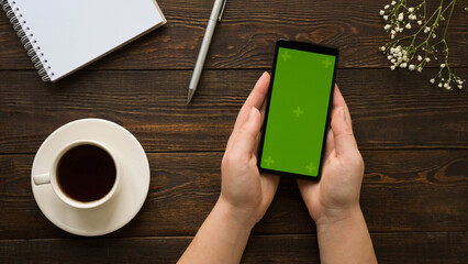 Composition with a hand holding a smartphone with a green screen, clean note, pen, a cup of coffee,...