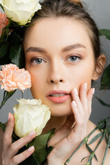 portrait of pretty woman with perfect skin touching face near roses and carnations isolated on grey.