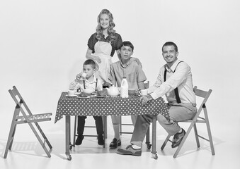 Portrait of lovely young family, woman, man and two boys sitting at the table and having breakfast isolated over grey background. Morning talk