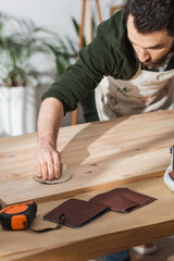 Bearded craftsman sanding wooden board near ruler in workshop.