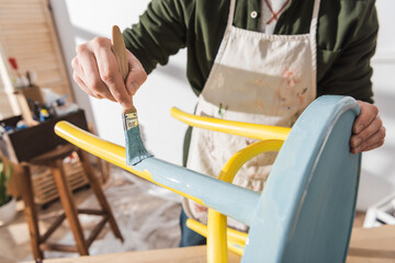 Cropped view of craftsman painting on wooden chair in blurred workshop.