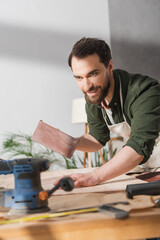 Positive restorer holding sandpaper near wooden board and sanding machine in workshop.