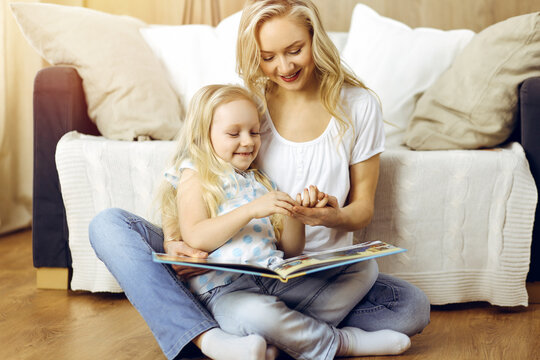 Happy family. Blonde young mother reading a book to her cute daughter while sitting at wooden floor in sunny room. Motherhood concept