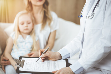 Fototapeta premium Doctor and patient. Pediatrician using clipboard while examining little girl with her mother at home. Sick and unhappy child at medical exam