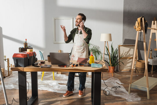 Cheerful carpenter talking on smartphone near tools on working table in studio.