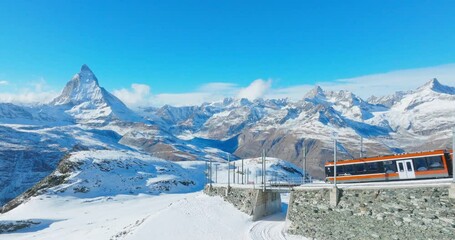Aerial drone footage of Gornergrat bahn railway running towards summit station with Matterhorn mountain peak background in Zermatt on a sunny winter day. Swiss Alps, Switzerland travel journey trip.