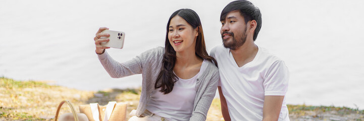  In love asian couple taking selfie photo with smartphone relaxing together with picnic Basket