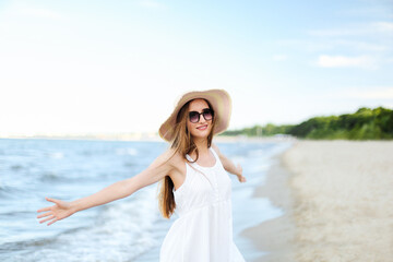 Fototapeta na wymiar Happy smiling woman in free happiness bliss on ocean beach standing with a hat, sunglasses, and open hands. Portrait of a multicultural female model in white summer dress enjoying nature 