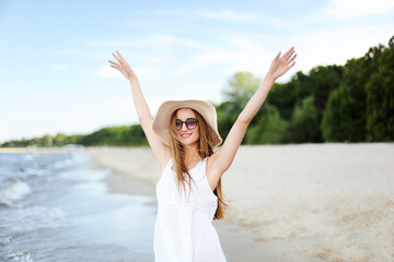 Fototapeta na wymiar Happy smiling woman in free happiness bliss on ocean beach standing with a hat, sunglasses, and raising hands. Portrait of a multicultural female model in white summer dress enjoying nature