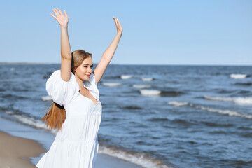 Fototapeta na wymiar Happy smiling beautiful woman on the ocean beach standing in a white summer dress, raising hands