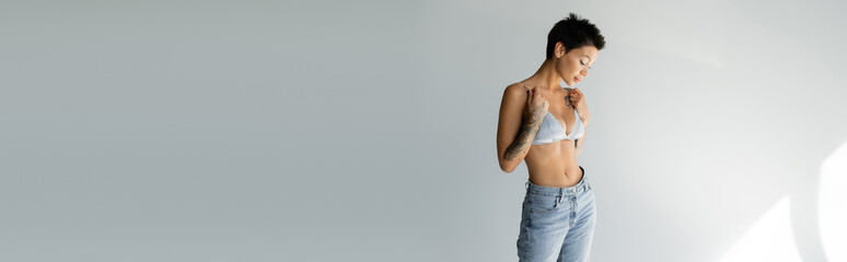 slender tattooed woman in blue jeans touching straps of silk bralette on grey background, banner.