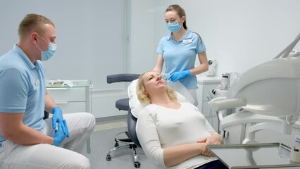 waiting for dental procedures a woman lies in chair man doctor and nurse stand next to thinking looking to side wearing masks gloves cleanliness and hygiene sterility filling prosthetics need doctor