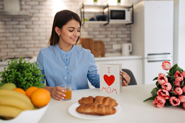 Happy mother's day! Attractive woman with flowers and postcard sitting on kitchen. Family holiday