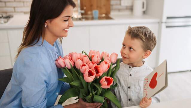 Young woman with her little son and greeting card for Mother's Day at home. Happy woman receiving flowers and greeting card from her son at home. Mother's day celebration
