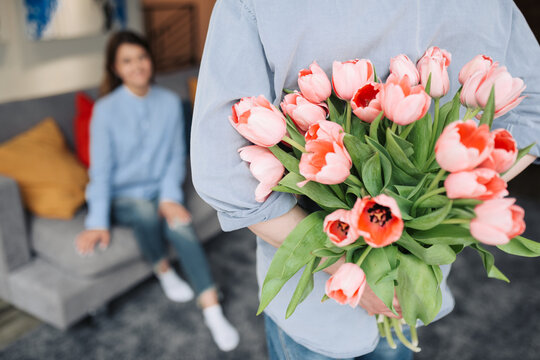 Unexpected moment in routine everyday life! Cropped photo of man's hands hiding holding chic bouquet of tulips behind back, happy woman is on background.