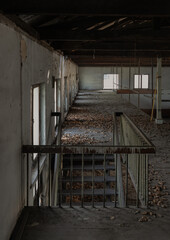 The old wooden stairs leading up to the mezzanine of an abandoned old building was left to deteriorate over time. Empty window frames and dried leaves filling the corridor. Space for text.