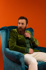 Fashionable bearded model sitting on velvet armchair on red background.