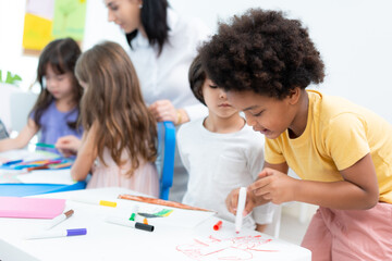 Selective focus of a little African American boy with Afro hair, standing drawing on paper with a red brush pen at a table next to a friend who sitting and watching while drawing at a kindergarten.