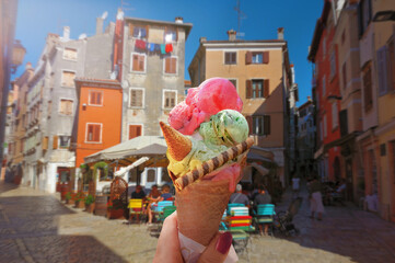Woman hand hold  beautiful bright  sweet ice - cream cone with different flavors   held in hand on the background of  old street  in  Rovinj, Croatia - 572726651