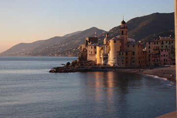 Fototapeta na wymiar Camogli, Italy - January 27, 2023: Beautiful old mediterranean town at the sunset time with illumination during winter days. People enjoying the evening at the beach with beautiful sunset background