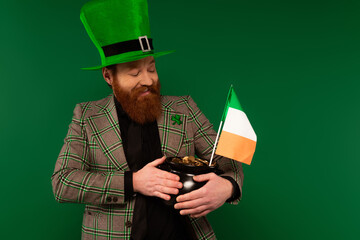 Positive man in hat holding pot with coins and Irish flag isolated on green.