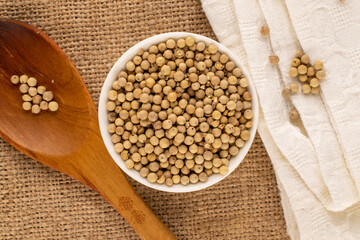 Fragrant vegetable coriander seeds in white plate with wooden spoon and white napkin on jute cloth, macro, top view.
