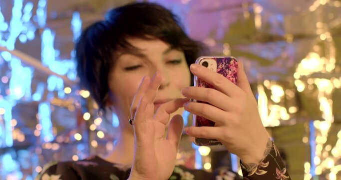 She uses the phone in her hands against the backdrop of light reflections. Check notifications and messengers at the party. Focus on the screen, press the finger, scroll.