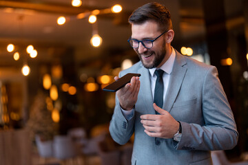 Handsome young businessman, restaurant manager wearing eyeglasses and business suit having amusing conversation on smart phone, laughing, talking on speakerphone while standing in modern restaurant.