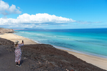 Mujer rubia con sombrero de paja y vestido de flores tomando una foto desde el acantilado en la playa de arena blanca y el mar turquesa de Jandia. Fuerteventura, Islas Canarias. 