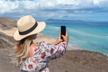 Chica joven rubia con un sombrero de paja tomando una foto con su teléfono móvil desde la colina rocosa hasta el mar turquesa y la playa blanca de Jandia en la turística Fuerteventura en las Islas Can