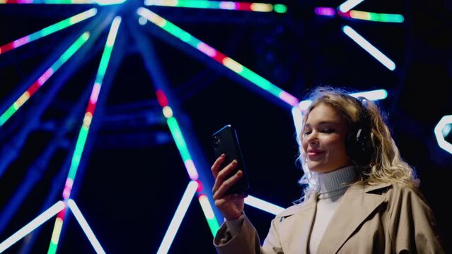 Carefree millennial female in headphones enjoying music playlist while standing near colorful illuminating ferris wheel.Young woman uses smartphone app for social media chat while reading text message